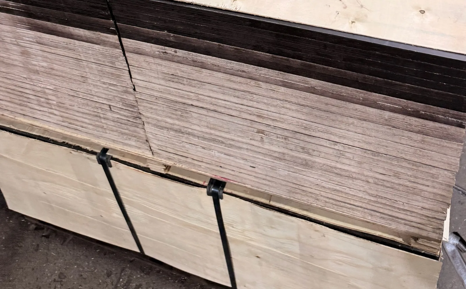 Corner of a strapped plywood bundle with steel bands and buckles over the layered sheets